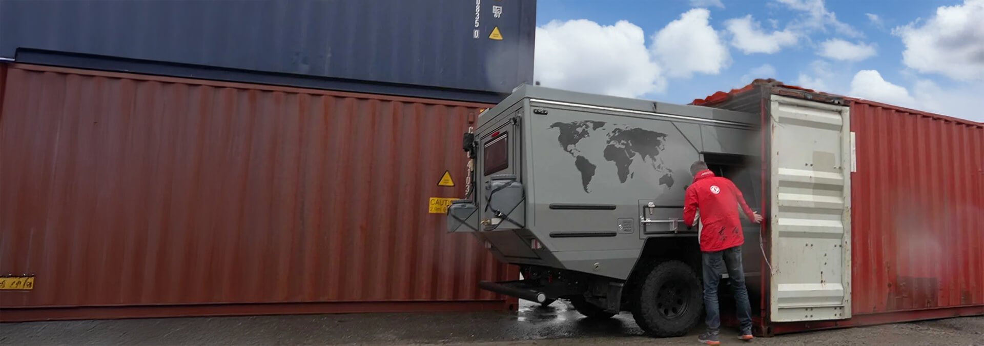 Ein Mann öffnet die Tür eines Wohnmobils neben Containern in einem Hafen. Wolken am Himmel sind sichtbar.